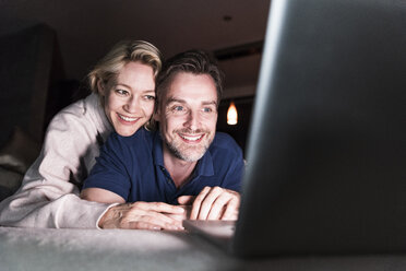 Glückliches Paar liegt auf der Couch zu Hause und schaut auf den Laptop - UUF13484