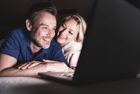 Glückliches Paar liegt auf der Couch zu Hause und benutzt einen Laptop - UUF13485