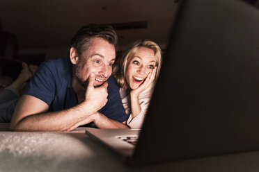 Erstauntes reifes Paar liegt zu Hause auf der Couch und schaut auf den Laptop - UUF13486