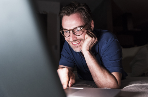 Porträt eines lächelnden Mannes, der zu Hause auf der Couch liegt und einen Laptop benutzt, lizenzfreies Stockfoto