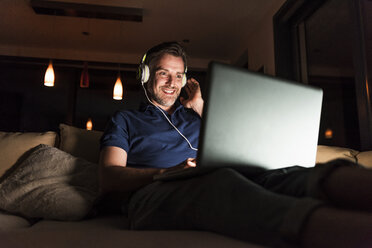 Man with headphones sitting on couch at home looking at laptop - UUF13492