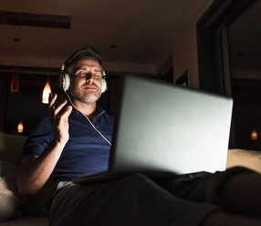 Mann sitzt auf der Couch zu Hause und hört Musik mit Kopfhörern und Laptop - UUF13493