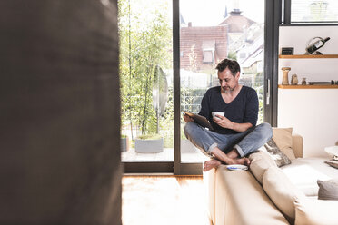 Mature man with cup of coffee sitting on backrest of couch using tablet - UUF13500