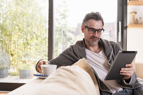 Porträt eines reifen Mannes mit einer Tasse Kaffee auf der Couch sitzend, der ein Tablet benutzt - UUF13501