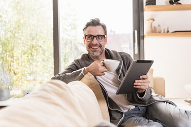 Porträt eines lächelnden Mannes mit einer Tasse Kaffee auf der Couch sitzend mit einem Tablet - UUF13502