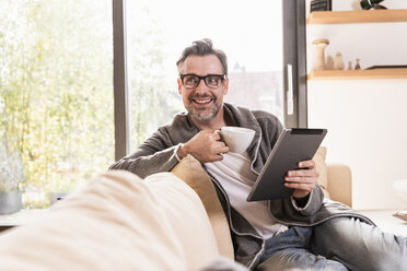 Portrait of smiling man with cup of coffee sitting on couch with tablet - UUF13502