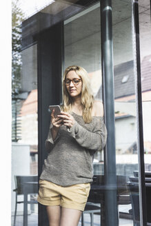 Porträt einer blonden reifen Frau, die vor einem Haus steht und auf ihr Smartphone schaut - UUF13518