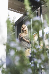 Content mature woman with coffee mug standing in front of her house - UUF13519