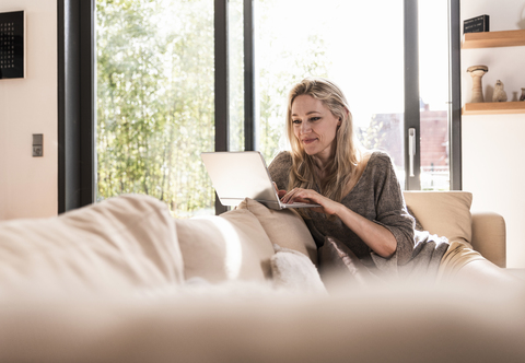 Lächelnde reife Frau sitzt zu Hause auf der Couch und benutzt einen Laptop, lizenzfreies Stockfoto