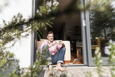 Content man sitting with cup of coffee at open terrace door - UUF13524