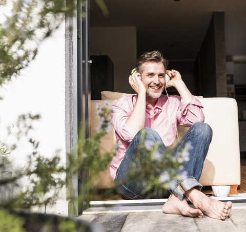 Lächelnder reifer Mann, der an der offenen Terrassentür sitzt und mit Kopfhörern Musik hört, lizenzfreies Stockfoto