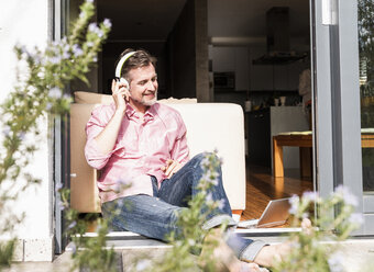 Smiling mature man sitting at open terrace door listening music with headphones - UUF13529