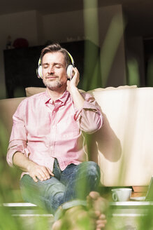 Porträt eines reifen Mannes, der an der offenen Terrassentür sitzt und mit Kopfhörern Musik hört - UUF13530