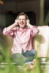 Porträt eines reifen Mannes, der an der offenen Terrassentür sitzt und mit Kopfhörern Musik hört - UUF13531
