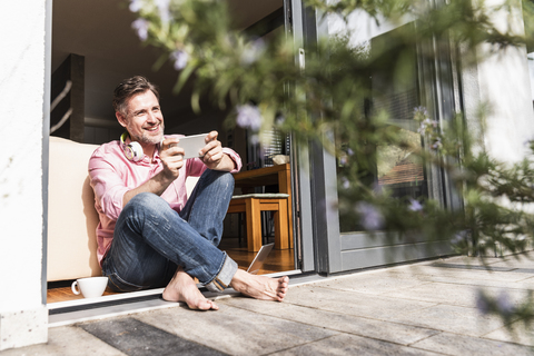 Lächelnder reifer Mann mit Smartphone sitzt an offener Terrassentür, lizenzfreies Stockfoto