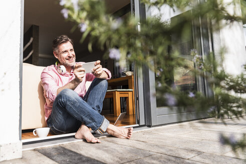Lächelnder reifer Mann mit Smartphone sitzt an offener Terrassentür - UUF13532