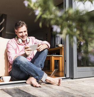 Lächelnder reifer Mann sitzt an offener Terrassentür und schaut auf sein Smartphone - UUF13533