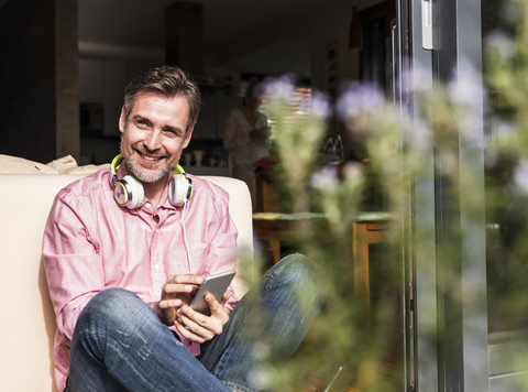 Porträt eines lächelnden reifen Mannes, der an der offenen Terrassentür sitzt und sein Smartphone benutzt, lizenzfreies Stockfoto