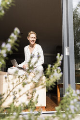 Lachende reife Frau mit Laptop an offener Terrassentür sitzend - UUF13538