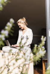 Lächelnde reife Frau sitzt an der offenen Terrassentür und benutzt einen Laptop - UUF13539