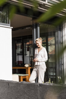 Lächelnde reife Frau steht auf der Terrasse eines Einfamilienhauses und schaut auf ihr Handy - UUF13542