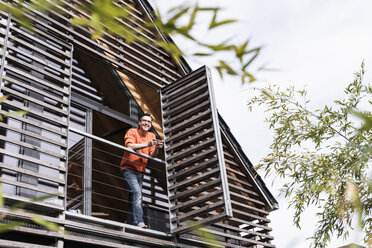 Man with smartphone standing on balcony of his house looking at distance - UUF13563