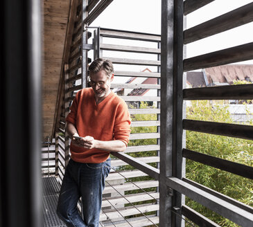 Lächelnder reifer Mann steht auf dem Balkon seines Hauses und schaut auf sein Smartphone - UUF13566