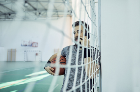 Mann mit Basketball und Smartphone hinter dem Netz, innen, lizenzfreies Stockfoto