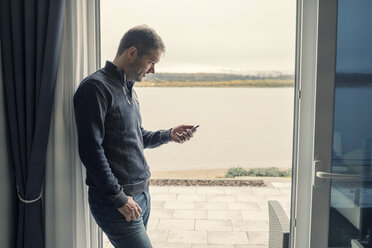 Mann steht in der offenen Tür seines Ferienhauses und liest Textnachrichten - PAF01828