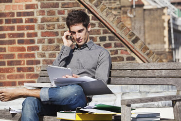 Junger Mann auf einer Bank sitzend mit einem digitalen Tablet und einem Mobiltelefon - ISF00479