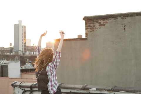 Young woman listening to music with arms raised on city rooftop stock photo