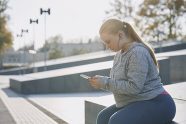 Kurvige junge Frau beim Training, an der Wand sitzend und auf das Smartphone schauend - CUF01961
