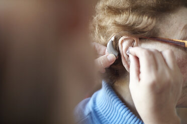 Mature woman helping senior woman insert hearing aid, close-up, differential focus - CUF02139