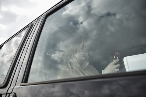 Junge Frau sitzt in einem Auto und schaut auf ihr Handy, lizenzfreies Stockfoto