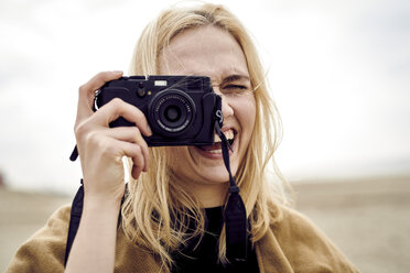Porträt einer blonden jungen Frau, die am Strand mit der Kamera fotografiert - MMIF00017