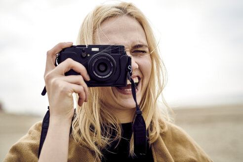 Porträt einer blonden jungen Frau, die am Strand mit der Kamera fotografiert - MMIF00017