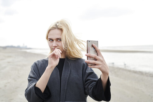 Netherlands, portrait of blond young woman taking selfie with smartphone on the beach - MMIF00021