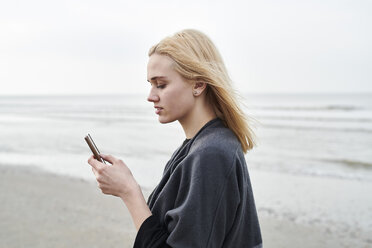 Niederlande, blonde junge Frau mit Smartphone am Strand - MMIF00023