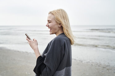 Niederlande, lachende blonde junge Frau mit Smartphone am Strand - MMIF00024