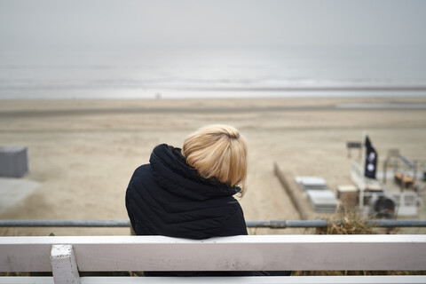 Back view of blond young woman sitting on bench looking to beach and sea in winter stock photo