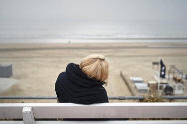 Rückenansicht einer blonden jungen Frau, die auf einer Bank sitzt und auf den Strand und das Meer im Winter schaut - MMIF00048