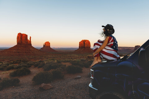 USA, Utah, Frau mit Flagge der Vereinigten Staaten von Amerika genießt den Sonnenuntergang im Monument Valley, lizenzfreies Stockfoto