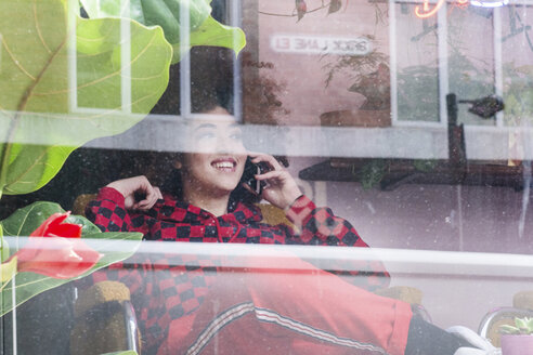 Junge Frau, die in einem Haus sitzt und ein Smartphone benutzt, gesehen durch ein Fenster - CUF02510