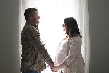 Pregnant woman holding hands with partner, face to face, in front of window - CUF02550