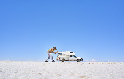 Falschperspektivisches Bild eines Jungen auf einer Salzwiese, der so tut, als würde er ein Freizeitfahrzeug schieben, Fahrzeug im Hintergrund, Salar de Uyuni, Uyuni, Oruro, Bolivien, Südamerika - CUF02607