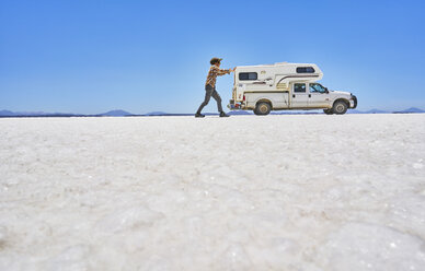 Falschperspektivisches Bild eines Jungen auf einer Salzwiese, der so tut, als würde er ein Freizeitfahrzeug schieben, Fahrzeug im Hintergrund, Salar de Uyuni, Uyuni, Oruro, Bolivien, Südamerika - CUF02609