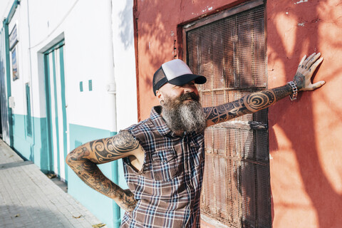 Tätowierter männlicher Hipster lehnt an einer sonnenbeschienenen Wand, lizenzfreies Stockfoto