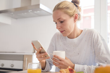 Junge Frau schaut am Frühstückstisch auf ihr Smartphone - CUF02750