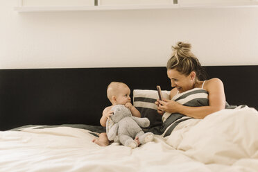 Mother photographing baby daughter sitting up in bed with soft toy - CUF02779