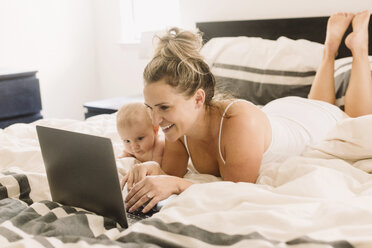 Baby-Mädchen und Mutter liegen auf dem Bett und schauen auf den Laptop - CUF02797